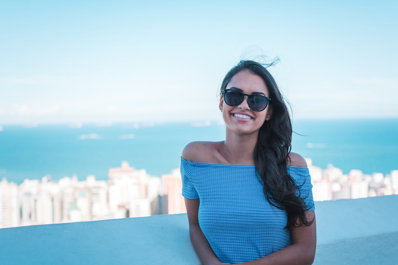 Portrait of a smiling woman wearing sunglasses, enjoying a sunny day with a cityscape background.